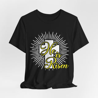 A black short-sleeve crewneck t-shirt with a graphic on the front featuring a white cross, white sunburst rays, and the text 'He is Risen' in yellow cursive script.