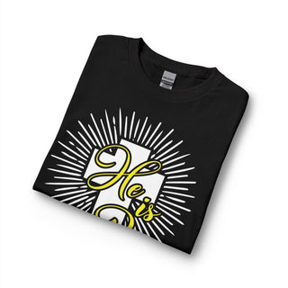 A folded black t-shirt with a graphic print featuring a white cross, white sunburst rays, and yellow cursive text reading 'He Is Risen displayed on a clean white background.
