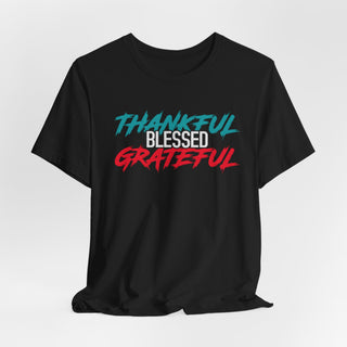 A folded black t-shirt with a graphic print reading 'THANKFUL BLESSED GRATEFUL' in teal, white, and red brushstroke-style lettering, shown on a plain white background.