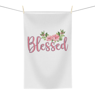 A white waffle-weave kitchen towel hanging from a clothesline with wooden clips, featuring the word 'Blessed' in pink script with a floral crown of pink flowers and green leaves above it.