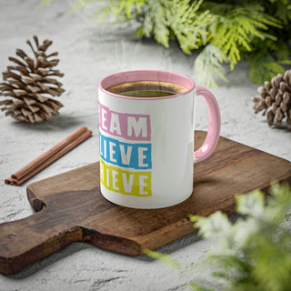 A white ceramic mug with a pink base and multicolored text that reads 'DREAM BELIEVE ACHIEVE in lifestyle background.